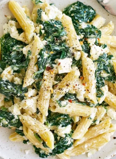 A plate of spinach feta pasta garnished with fresh spinach and feta cheese.